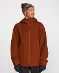A person wearing the Volcom Men’s Brighton Full Zip Jacket in rust, featuring a V-Science 2-Layer construction and PFAS-free DWR, along with black gloves and a patterned gray beanie, stands turned slightly to the side and looks off-camera against a plain light background.