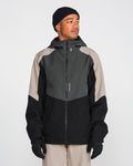 A person wearing the Volcom Men’s Brighton Full Zip Jacket—a black and gray hooded jacket with V-Science 2-Layer construction and a PFAS-free DWR finish—along with beige pants, black gloves, and a black beanie, stands in front of a plain white background, looking slightly to the side.