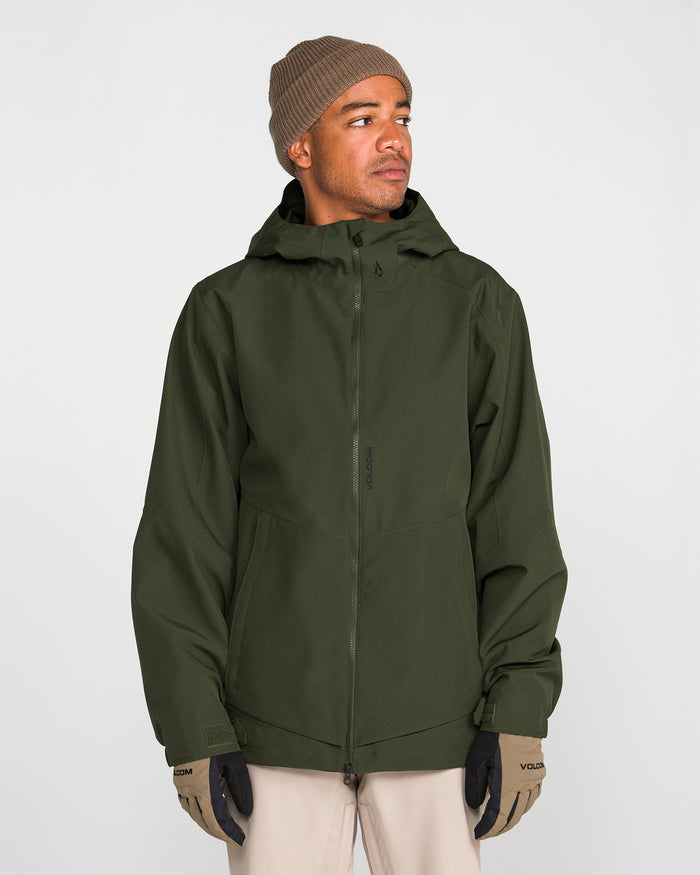 A person in a tan beanie, tan gloves, and beige pants stands facing slightly right against a plain light background, wearing the Volcom Men’s Brighton Full Zip Jacket—a green hooded jacket with fully taped seams.