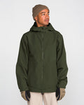 A person in a tan beanie, tan gloves, and beige pants stands facing slightly right against a plain light background, wearing the Volcom Men’s Brighton Full Zip Jacket—a green hooded jacket with fully taped seams.