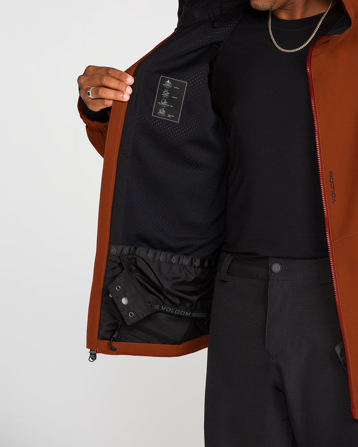 A person wearing black pants and a dark shirt displays the interior of the Volcom Men’s Brighton Full Zip Jacket, highlighting its black mesh lining, inner label, and innovative Zip Tech jacket-to-pant interface. The individual’s face is not visible.