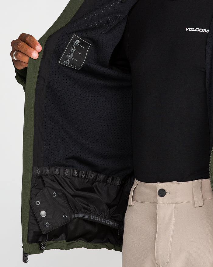 A person opens a green Volcom Men’s Brighton Full Zip Jacket to reveal its black mesh inner lining and interior tag, while wearing beige pants and a black VOLCOM shirt.