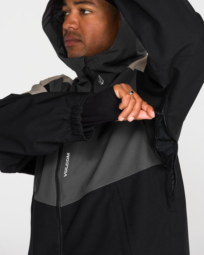 A person wearing the Volcom Men’s Brighton Full Zip Jacket in black and gray, featuring V-Science 2-Layer fabric, unzips a side vent to reveal a thumb hole cuff. Their face is partially visible against a plain white background.