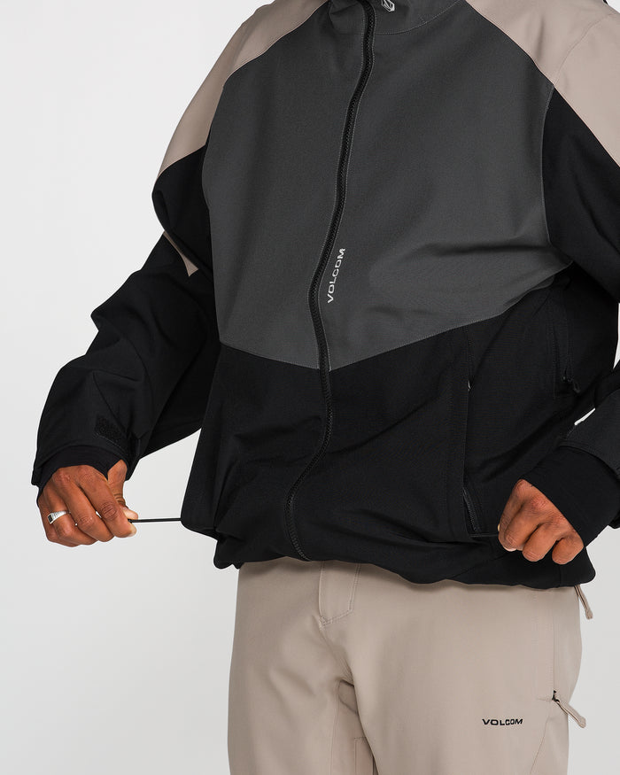 A person wearing the Volcom Men’s Brighton Full Zip Jacket with bold color-blocking and beige pants adjusts the drawstring at the hem. The jacket, displaying “VOLCOM” on both the zipper and chest, combines style and performance with a PFAS-free DWR finish. The person’s head is not visible.