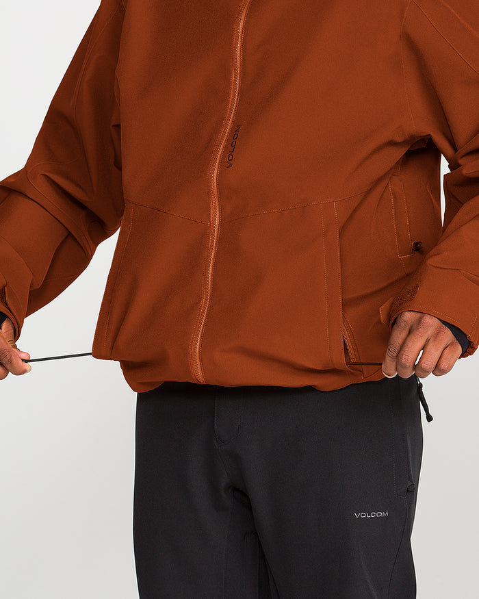 A person wearing Volcom’s Men’s Brighton Full Zip Jacket in rust and black pants adjusts the drawstring at the jacket’s hem; only their torso and hands are visible.
