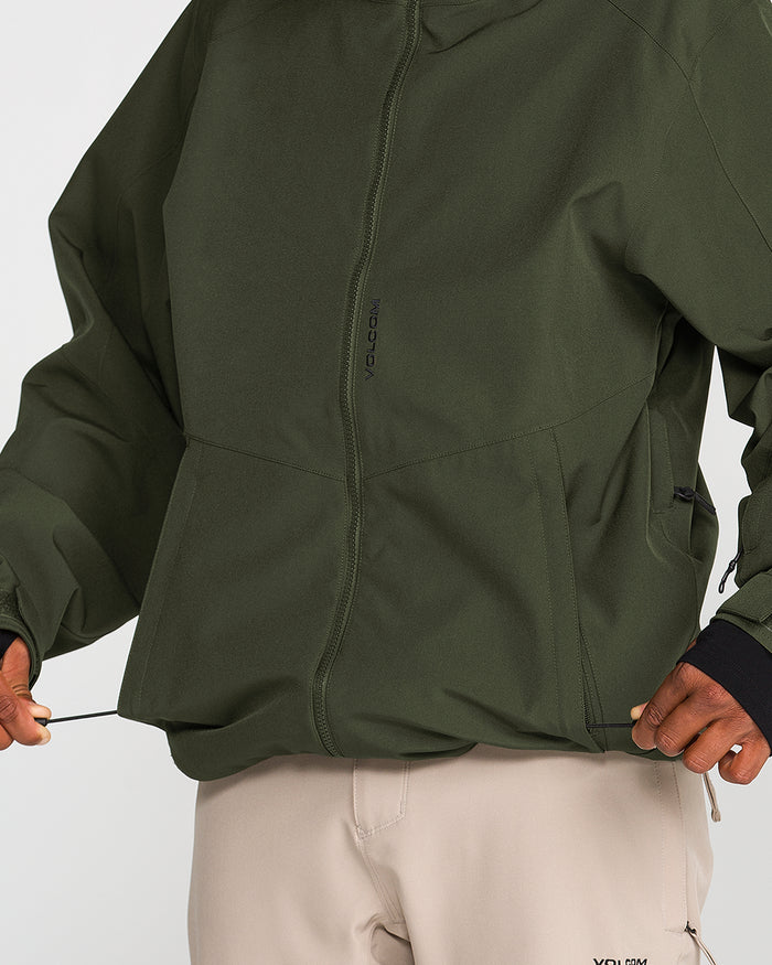 A person wearing the Volcom Men’s Brighton Full Zip Jacket in olive green and light beige pants adjusts the jacket’s drawstrings. The jacket, made from recycled stretch material, features subtle Volcom branding and a front zipper with a Zip Tech interface. Only the torso and hands are visible.