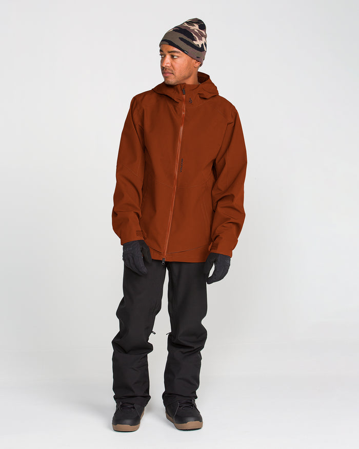 A person wearing the Volcom Men’s Brighton Full Zip Jacket in brown, which features V-Science 2-Layer construction and a Zip Tech jacket-to-pant interface, along with black pants, black gloves, black shoes, and a patterned beanie stands facing slightly to the side against a plain white background.