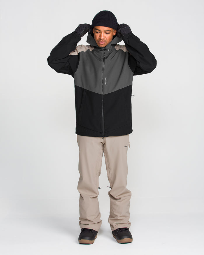 A person wearing the Volcom Men’s Brighton Full Zip Jacket in black and gray with PFAS-free DWR, paired with beige pants, black gloves, a black beanie, and dark shoes stands facing forward, adjusting their hood against a plain white background.