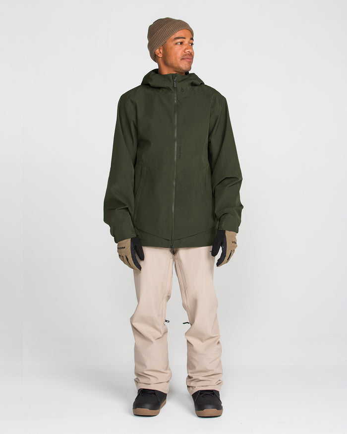 A person is standing against a plain white background, wearing the Volcom Men’s Brighton Full Zip Jacket in green recycled stretch fabric with fully taped seams, paired with beige pants, brown gloves, a brown beanie, and black shoes. The person is looking to the side and smiling.
