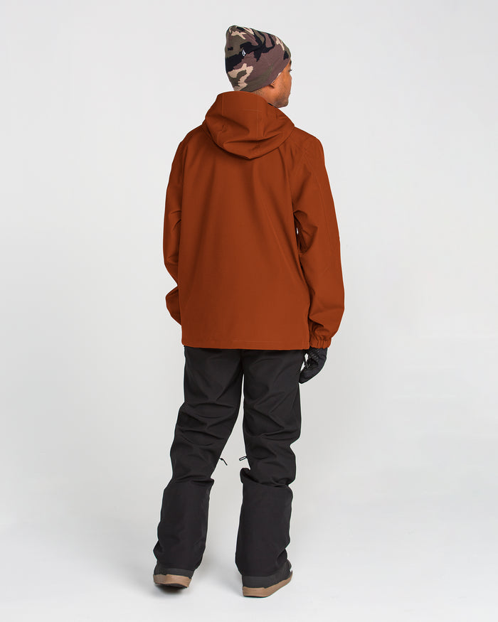 A person wearing a brown Volcom Men’s Brighton Full Zip Jacket with a hood, black pants featuring a Zip Tech jacket-to-pant interface, brown boots, black gloves, and a camouflage beanie stands facing away against a plain white background.