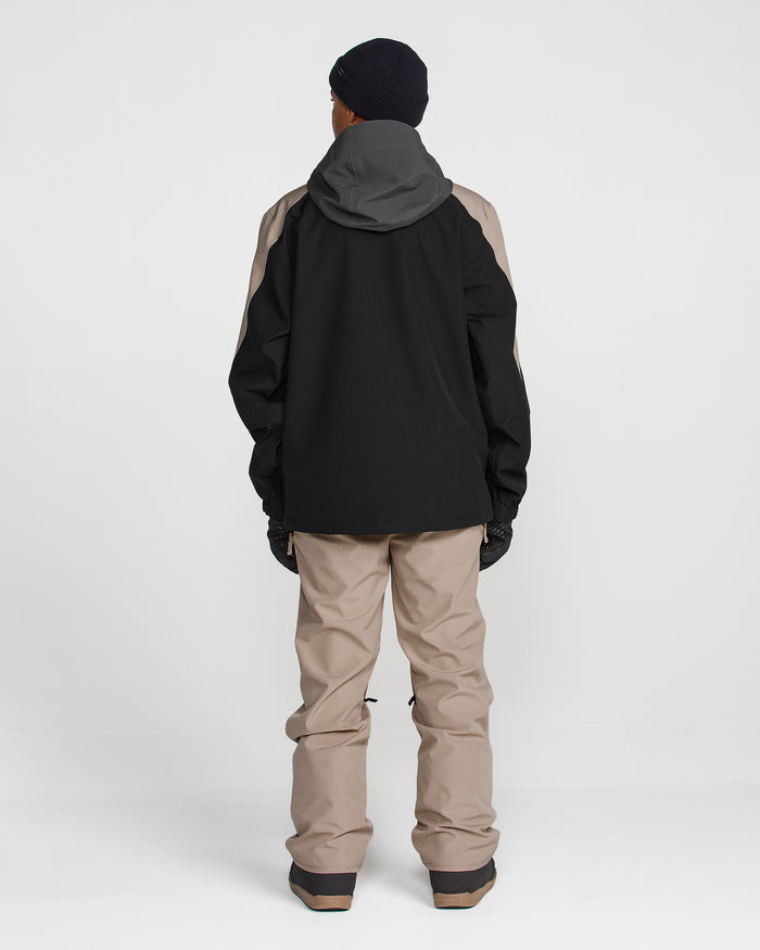 A person is standing facing away from the camera, wearing a black and gray Volcom Men’s Brighton Full Zip Jacket featuring a Zip Tech jacket-to-pant interface, beige pants, black gloves, black boots, and a black beanie, against a plain white background.