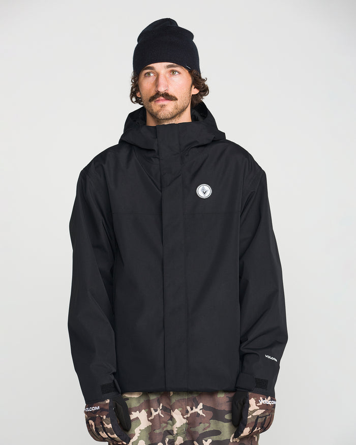 A man wearing the Volcom Men’s Buckthreeeighty Insulated Jacket made from recycled fabric, a black beanie, and camo pants stands facing forward with a neutral expression. He is also wearing black gloves and has a mustache. The background is plain and light-colored.