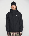 A man wearing the Volcom Men’s Buckthreeeighty Insulated Jacket made from recycled fabric, a black beanie, and camo pants stands facing forward with a neutral expression. He is also wearing black gloves and has a mustache. The background is plain and light-colored.