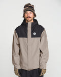 A man with a mustache wears the Volcom Men’s Buckthreeeighty Insulated Jacket, featuring a beige and black design made from recycled fabric, along with a patterned beanie and beige gloves. He stands against a plain white background, gazing slightly upward.