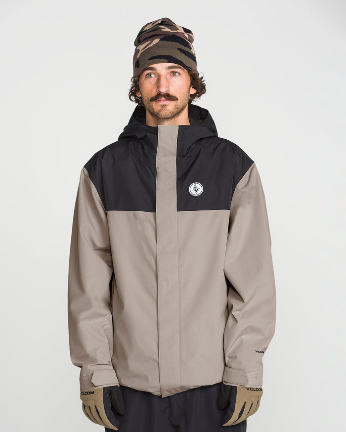 A man with a mustache wears the Volcom Men’s Buckthreeeighty Insulated Jacket, featuring a beige and black design made from recycled fabric, along with a patterned beanie and beige gloves. He stands against a plain white background, gazing slightly upward.