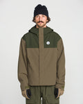 A man stands facing forward against a plain white background, wearing the Volcom Men’s Buckthreeeighty Insulated Jacket in two-tone green and olive—crafted from recycled fabric—paired with olive pants, a black beanie, and gloves.