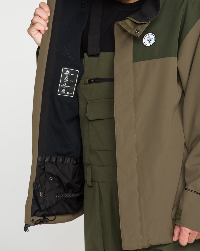 Wearing the Volcom Men’s Buckthreeeighty Insulated Jacket in two-tone green, a person holds it open to display the interior details and visible label, revealing coordinating green overalls underneath. Made from recycled fabric, this jacket by Volcom combines sustainability with standout style.