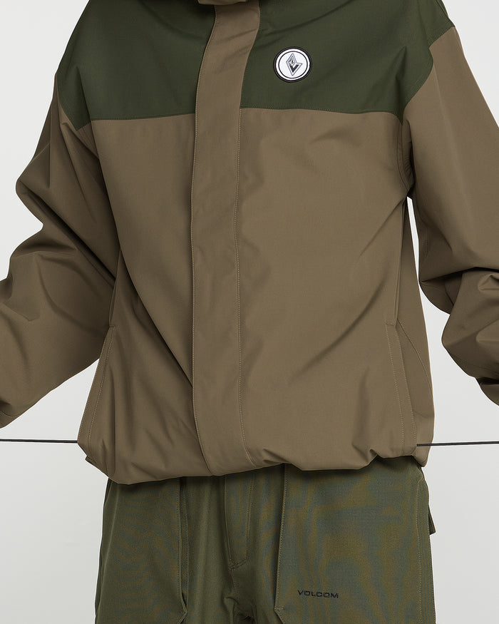 A person is seen wearing the Volcom Men’s Buckthreeeighty Insulated Jacket in green and brown, featuring a 10K waterproof rating, a hood, and a round chest patch, paired with matching pants. Their arms are slightly raised and their face is not visible.