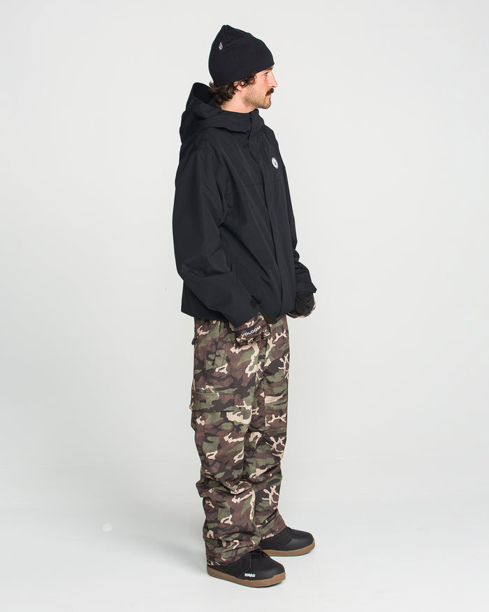A man stands sideways against a plain white background, wearing a black beanie, camo pants, black shoes, and the Volcom Men’s Buckthreeeighty Insulated Jacket made from recycled fabric.