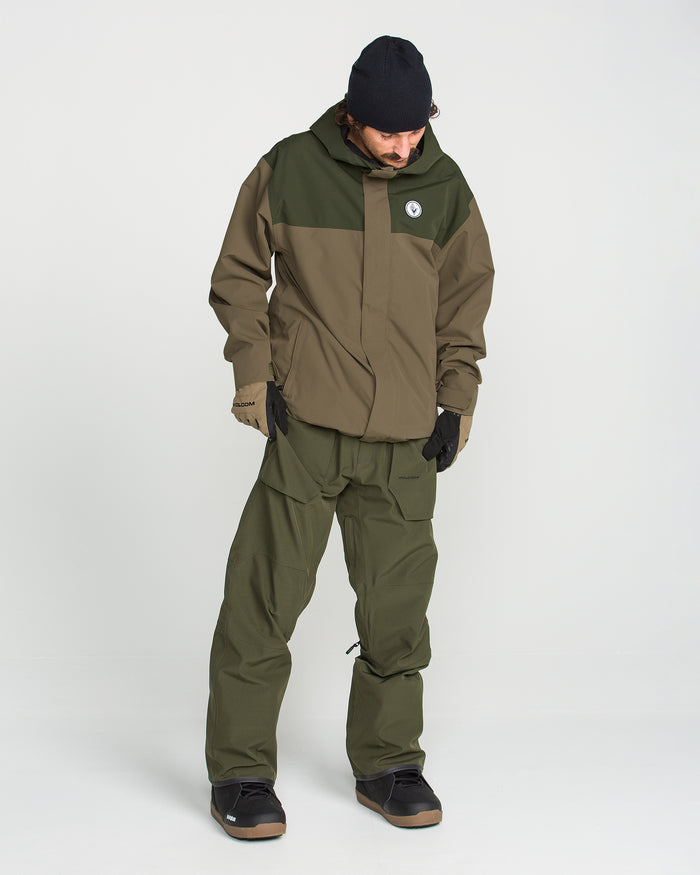 A person wearing the Volcom Men’s Buckthreeeighty Insulated Jacket in a two-tone olive and brown design, crafted from recycled fabric, paired with matching pants, black gloves, a black beanie, and black shoes stands against a plain white background looking down.