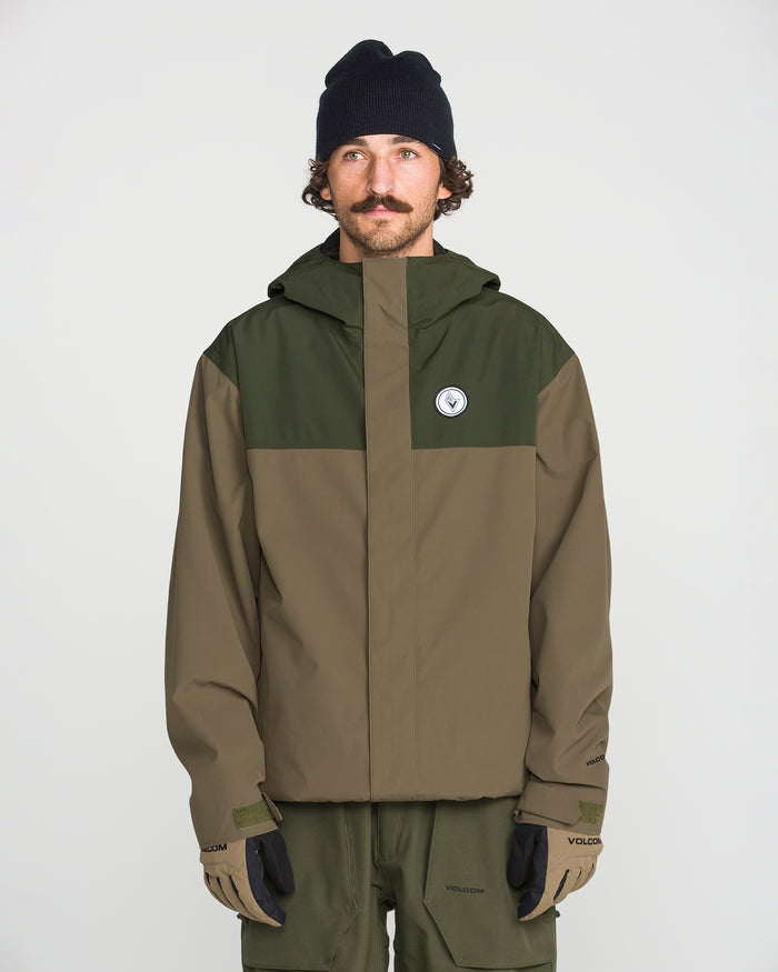 A person wearing the Volcom Men’s Buckthreeeighty Jacket in an olive green and brown baggy fit—crafted with V-Science 2-Layer fabric and PFAS-free DWR—paired with olive pants, a black beanie, and gloves, stands against a plain white background.