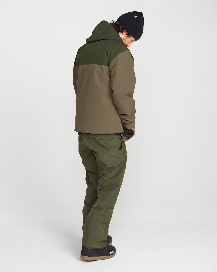 A person wears the Volcom Men’s Buckthreeeighty Jacket in olive green with a baggy fit, paired with matching pants, a black beanie, and black boots, standing against a plain white background with their head turned down. The jacket features a PFAS-free DWR finish for added protection.
