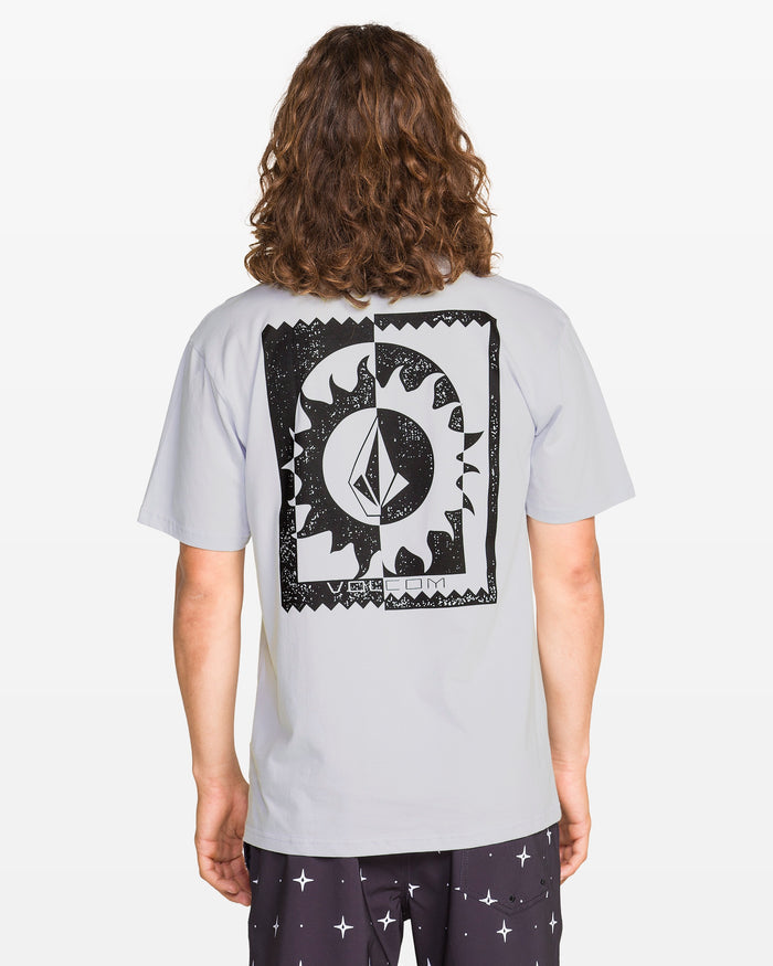 A person with long, wavy hair wears the Volcom Men's Burner Short Sleeve Tee in light gray, featuring a large black and white geometric sun and moon print on the back, paired with black shorts with a white star pattern.