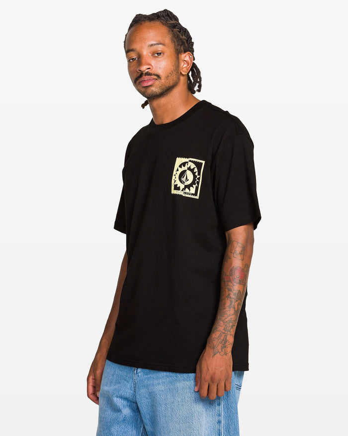 A man with braided hair and tattoos on his right arm wears the Volcom Men's Burner Short Sleeve Tee featuring a small chest graphic paired with light blue jeans, standing against a plain white background.
