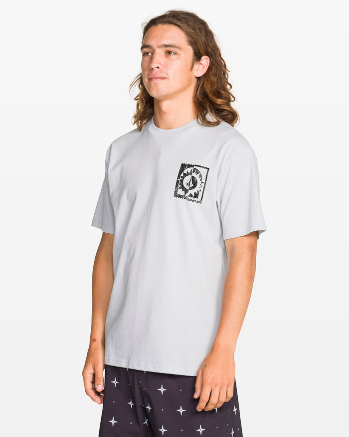 A person with long brown hair wears the Volcom Men's Burner Short Sleeve Tee with a black graphic soft-hand print on the left chest and dark shorts with a white cross pattern, standing against a plain white background.