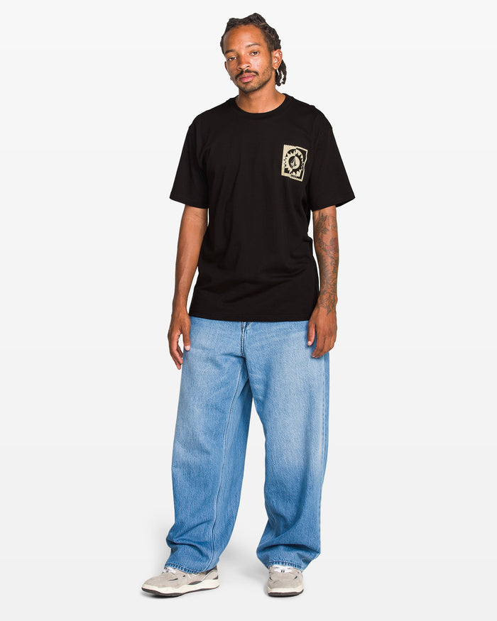 A man wears the Volcom Men's Burner Short Sleeve Tee, featuring a beige chest pocket graphic, paired with light blue baggy jeans and white sneakers, standing against a plain white background.