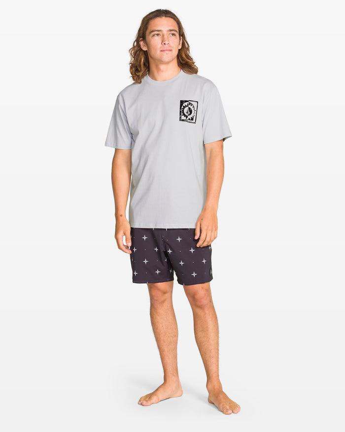 A person with long brown hair stands barefoot against a white background, wearing Volcom’s Men's Burner Short Sleeve Tee in light gray and black shorts with white star patterns.