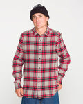 A person wearing the Volcom Men's Caden Plaid Long Sleeve Shirt stands against a plain white background, paired with blue jeans and a black beanie, looking at the camera with a neutral expression. The shirt’s classic fit highlights its timeless style.