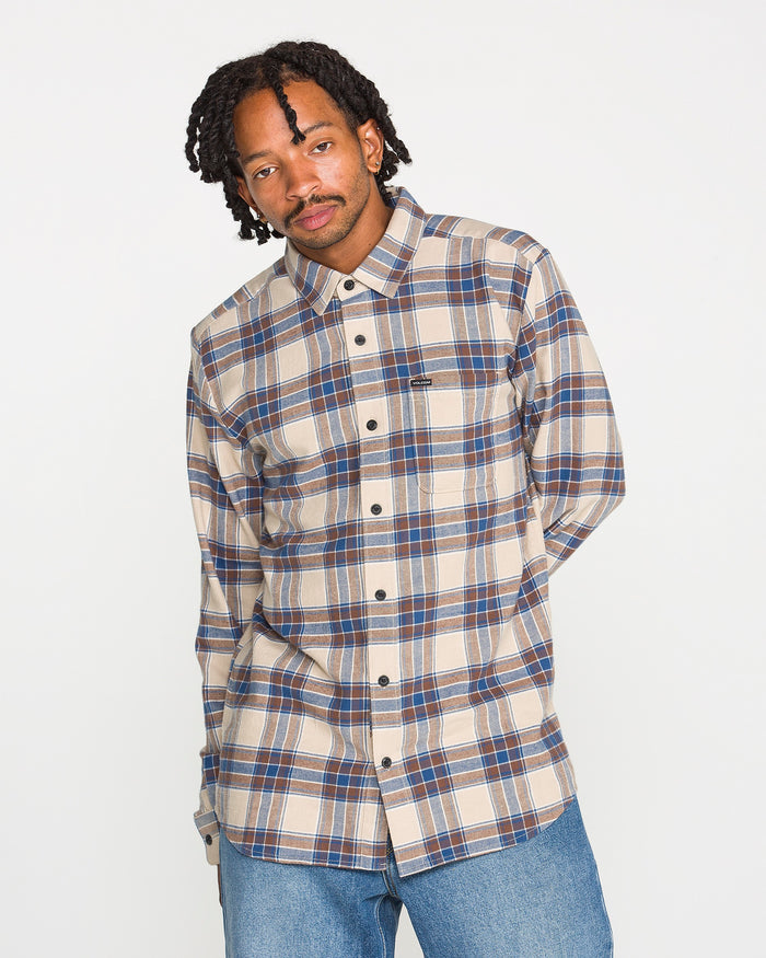 A man with short dreadlocks wearing the Volcom Men's Caden Plaid Long Sleeve Shirt in a classic fit, paired with light blue jeans, stands against a plain white background.