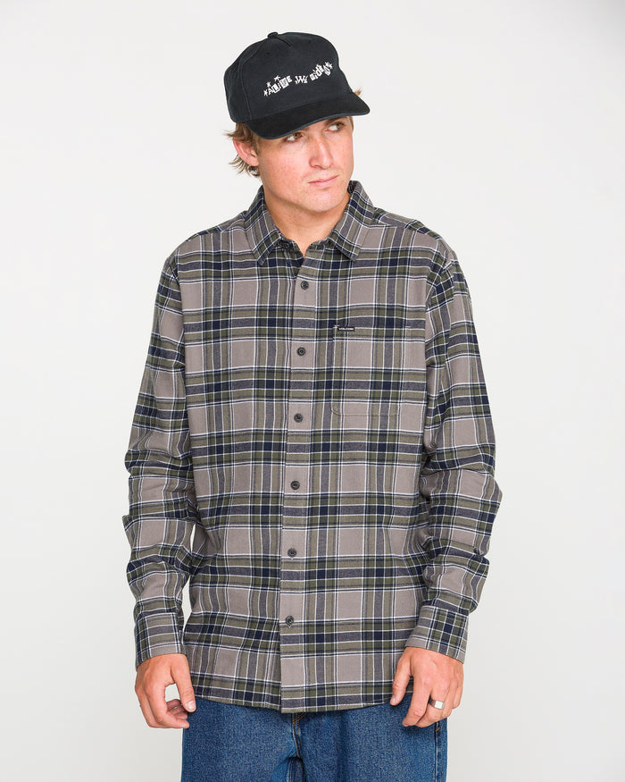 A man models the Volcom Men's Caden Plaid Long Sleeve Shirt in classic fit gray and dark blue, paired with a black cap and blue jeans, standing against a plain white background and looking slightly to the side.