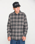 A man models the Volcom Men's Caden Plaid Long Sleeve Shirt in classic fit gray and dark blue, paired with a black cap and blue jeans, standing against a plain white background and looking slightly to the side.