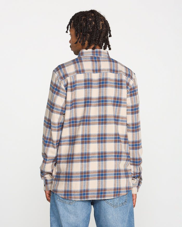 A person with short dreadlocks is shown from the back, wearing the Volcom Men's Caden Plaid Long Sleeve Shirt in beige, blue, and brown, paired with light blue jeans against a plain white background.