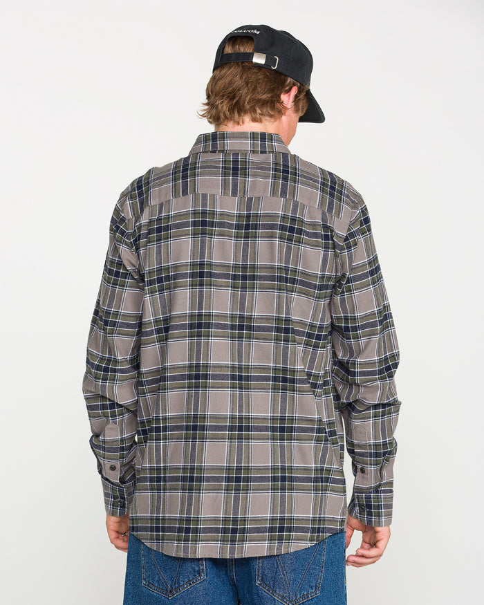 A person is standing with their back to the camera against a plain white background, wearing a black cap, blue jeans, and the Volcom Men's Caden Plaid Long Sleeve Shirt in a classic fit gray and blue plaid.