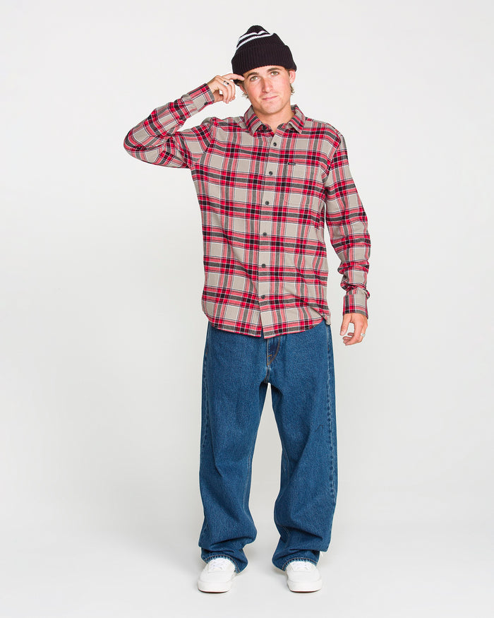 A person wearing a black beanie, blue jeans, white sneakers, and the Volcom Men's Caden Plaid Long Sleeve Shirt in red stands against a plain white background, touching the brim of their beanie with one hand.