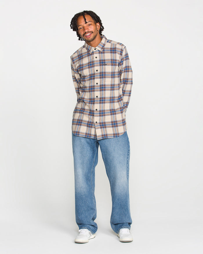 A man wearing the Volcom Men's Caden Plaid Long Sleeve Shirt in a classic beige and blue pattern, paired with loose blue jeans and white sneakers, stands against a plain white background, smiling with his hands in his pockets.