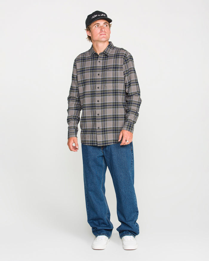 A man wearing a black cap, blue jeans, and white sneakers stands against a plain white background, looking slightly to his left. He is dressed in the Volcom Men's Caden Plaid Long Sleeve Shirt in a classic fit.