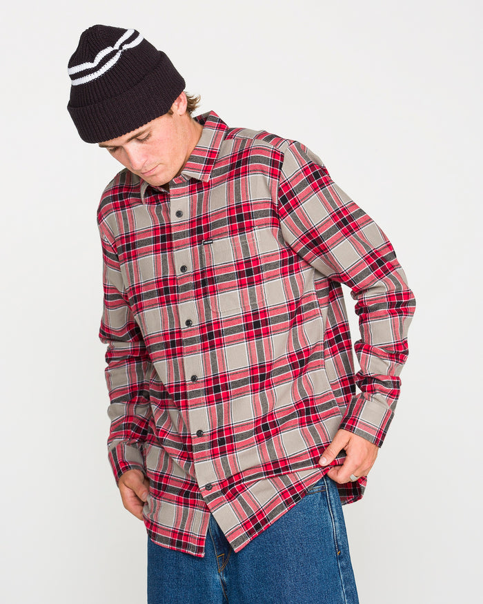 A person wearing the Volcom Men's Caden Plaid Long Sleeve Shirt in red and gray with classic fit, paired with blue jeans and a black knit beanie, looks down while adjusting the shirt hem against a plain white background.