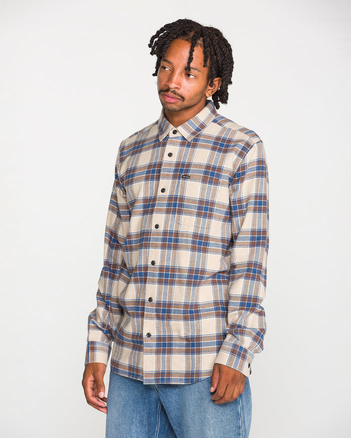 A man with short dreadlocks wears the Volcom Men's Caden Plaid Long Sleeve Shirt, featuring a classic fit in beige, blue, and brown plaid, paired with light blue jeans. Crafted from soft cotton flannel, the shirt stands out as he poses against a plain white background, looking slightly to his left with a neutral expression.