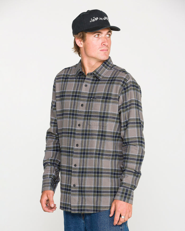 A man wearing a black baseball cap, jeans, and the Volcom Men's Caden Plaid Long Sleeve Shirt stands against a plain white background, looking to his left.