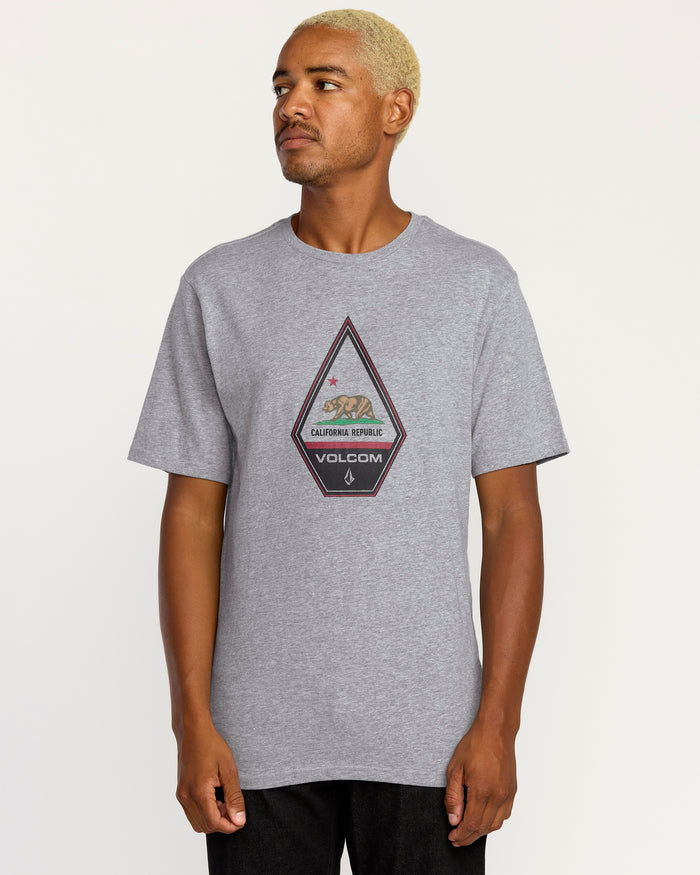 A person with short blond hair wears a relaxed fit gray Volcom Men's Californeye Tee featuring a triangular graphic, standing against a plain white background.