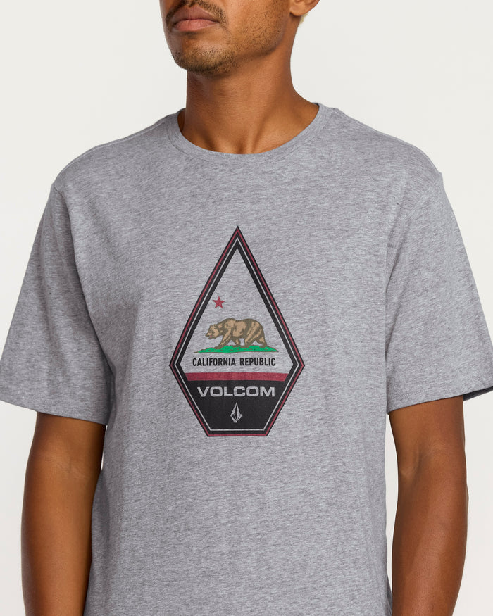 A person wears the Volcom Men's Californeye Tee—a light gray, relaxed fit heathered shirt with a chest patch showing a bear, red star, "California Republic," and Volcom branding. The partially visible face adds graphic edge to the look.