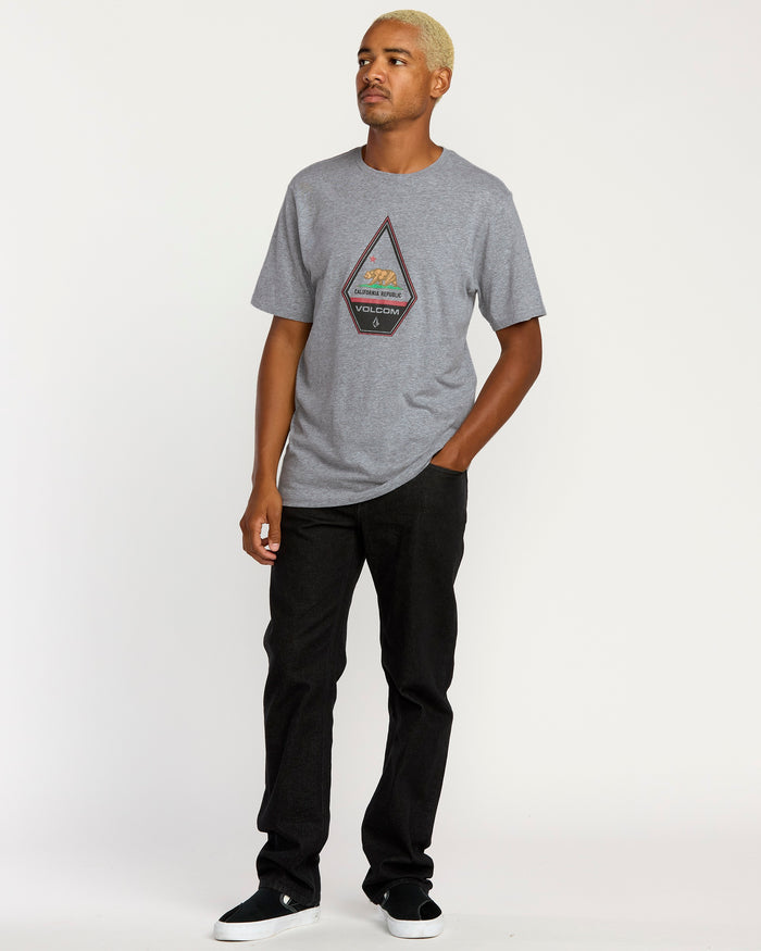 A man with short blond hair wears the Volcom Men's Californeye Tee in gray heather, black pants, and black slip-on shoes, posing against a plain white background with one hand in his pocket for a relaxed look.