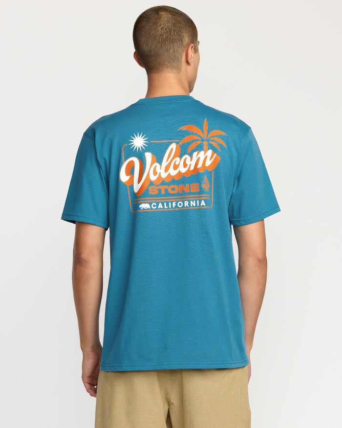 A person, seen from behind, is wearing the Volcom Men's California Dreaming Short Sleeve Tee in blue, which features an orange and white palm tree, sun, and "California" graphic on the back.