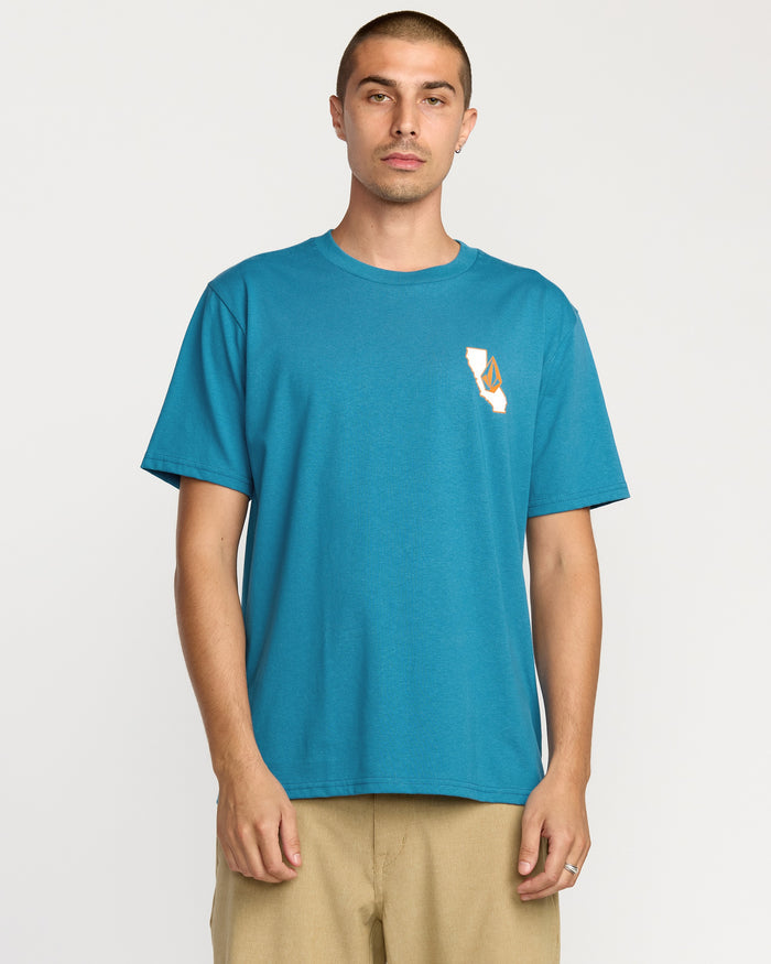 A man with short hair wears the Volcom Men's California Dreaming Short Sleeve Tee in teal and beige pants, standing against a white background. The classic fit tee features a small soft-hand screen print graphic on the left chest.