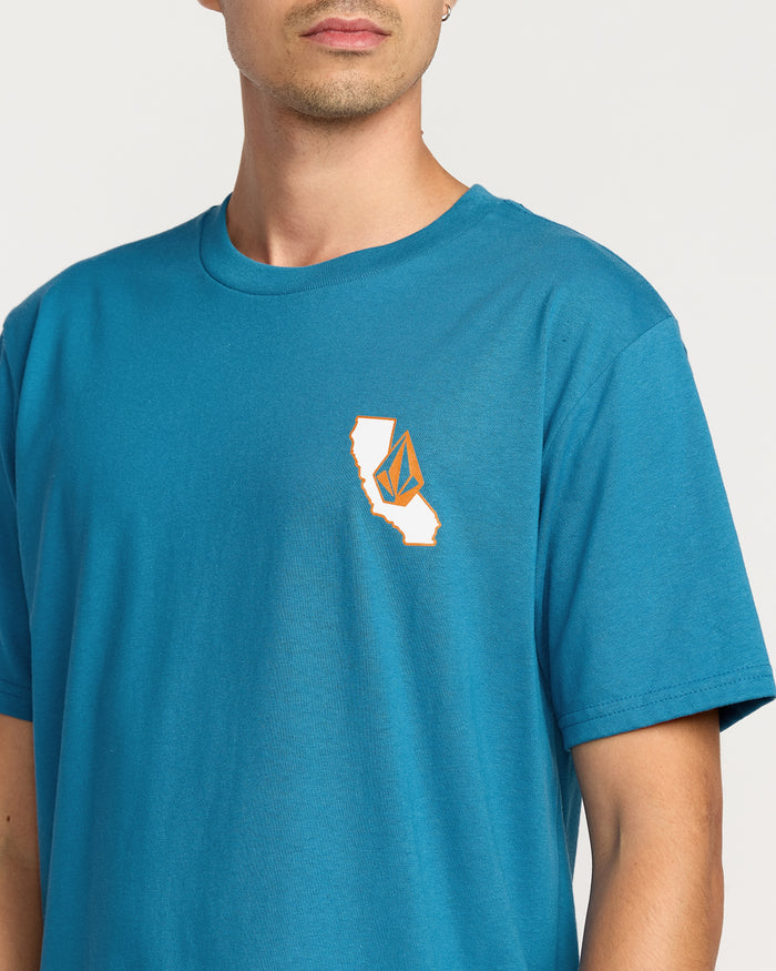 A person wears the Volcom Men's California Dreaming Short Sleeve Tee in blue, a classic fit with a small California graphic and abstract logo on the left chest, featuring a soft-hand screen print on premium cotton.