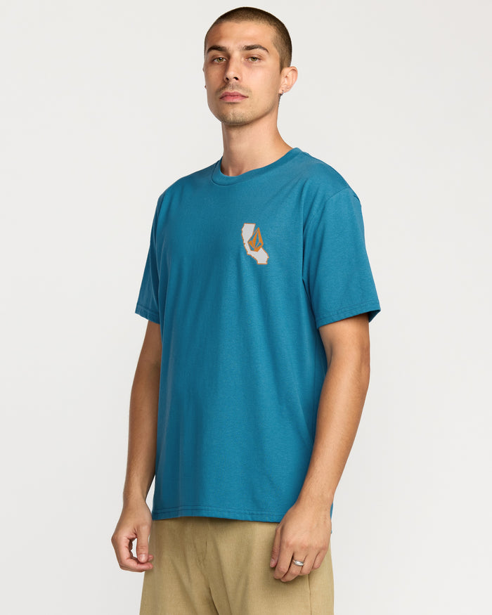 A man wears the Volcom Men's California Dreaming Short Sleeve Tee in teal, featuring a soft-hand screen print on the chest, paired with beige pants, standing against a plain white background.